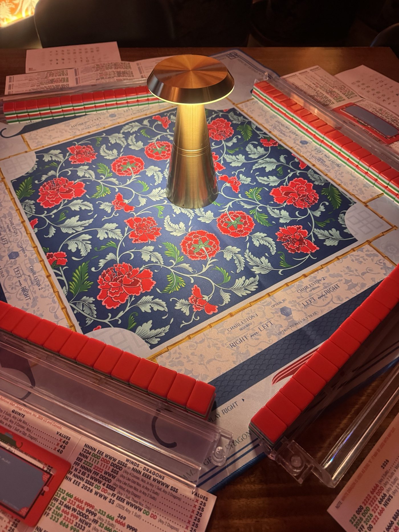 A mahjong set laid out on a blue and red peony mat, lit by a gold table lamp